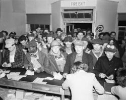 一大群工人擠在櫃檯前，兩名女櫃員正在俯身寫字。有的工人將自己的證件照放在帽沿上戴着。