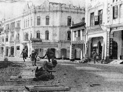 Japanese troops running along a rubble road in front of old colonial buildings.