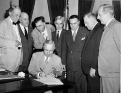 一名身着西裝的男子坐在桌子旁，正在簽署一份文件。他周圍站着另外七名身着西裝的男子。