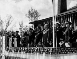 一群男性身着西装或军装，站在一个彩旗飘扬的舞台上，有的在敬礼。